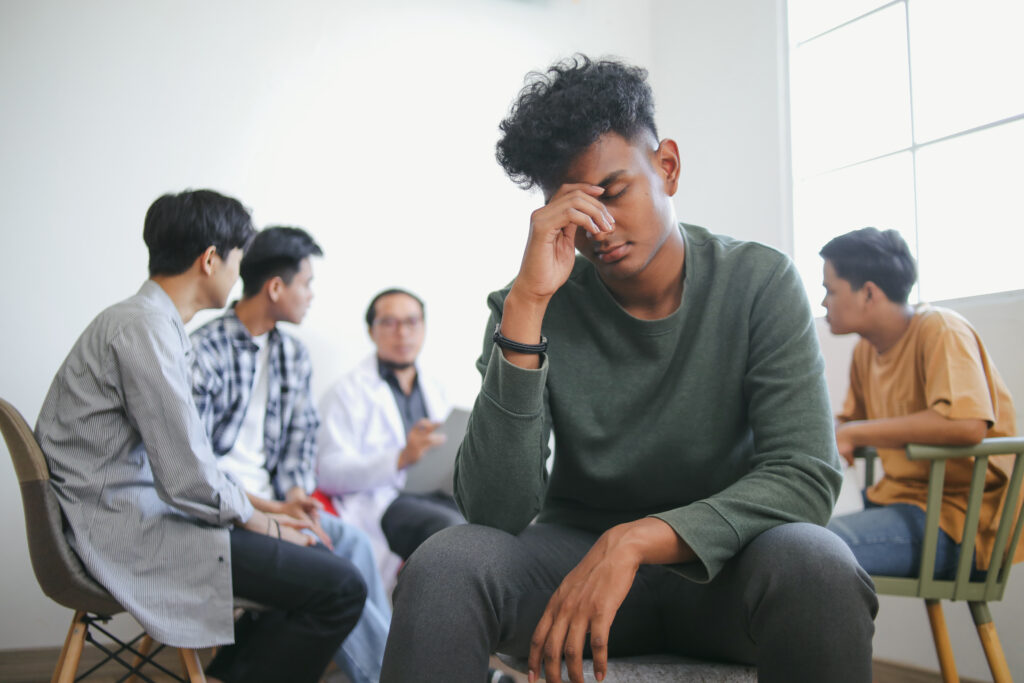 stress vs anxiety - Portrait Of Stressed Young Indian Guy Against Blurry Group Therapy Session Background